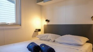 Ein ordentlich bezogenes Bett mit weißen Laken und zwei dunklen Handtüchern mit einem Logo, eine Nachttischlampe und ein Fenster mit Jalousien.