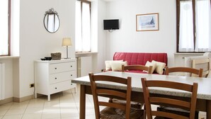 Een eetgedeelte met een tafel, stoelen, een wit kastje, een spiegel, een televisietoestel en een raam met gordijnen.
