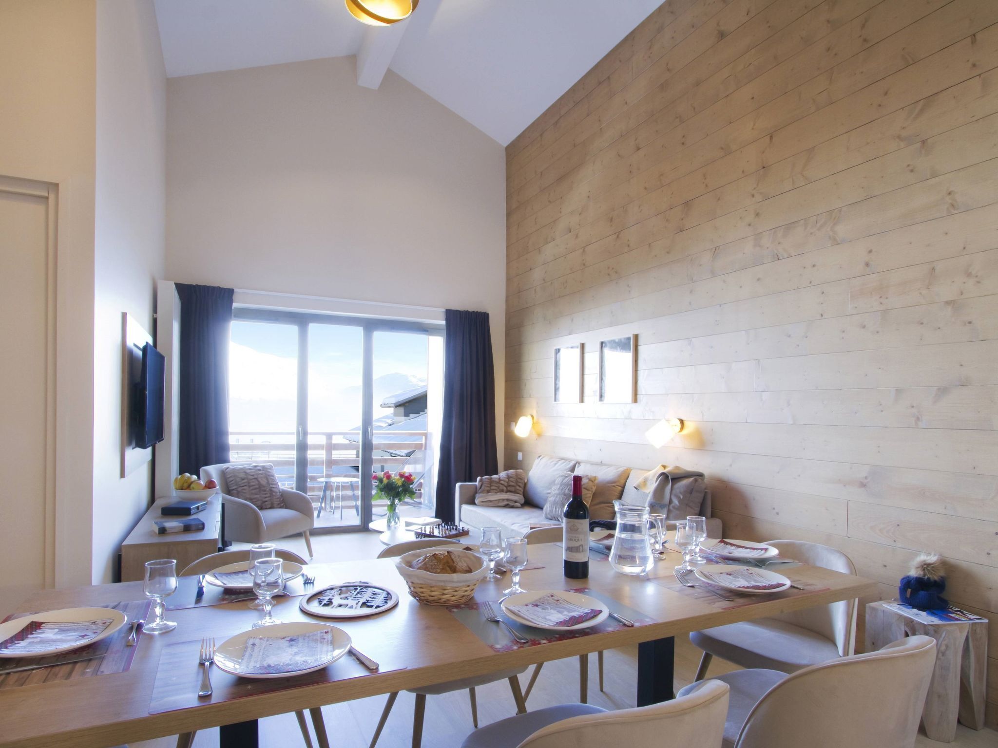 A dining area with a large table set for a meal, a television mounted on the wall, and a view of the outdoors through a sliding glass door.