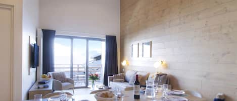 Uma sala de jantar com uma mesa grande posta para refeição, uma televisão fixada na parede e vista para o exterior por uma porta de correr de vidro.