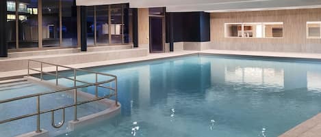 An indoor swimming pool with clear water, a metal safety railing, and a modern interior design with large windows and wooden accents.