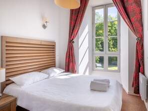 A bedroom with a wooden headboard, white bedding, a window with red curtains, and a lamp on the nightstand.