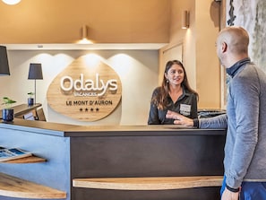 Un mostrador de recepción con un letrero de madera que dice 'Odalys Vacances Le Mont d'Auron' y una tarjeta de identificación para un miembro del personal.
