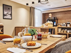Una escena de desayuno con una mesa dispuesta para dos, que incluye un croissant, un vaso de zumo de naranja, un bol y un tarro de mermelada.