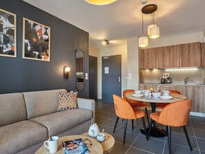 Ein modernes Wohnzimmer mit einer Couch, einem Couchtisch und orangefarbenen Sesseln. Im Hintergrund ist eine Küche mit Holzausbau und Spüle zu sehen.