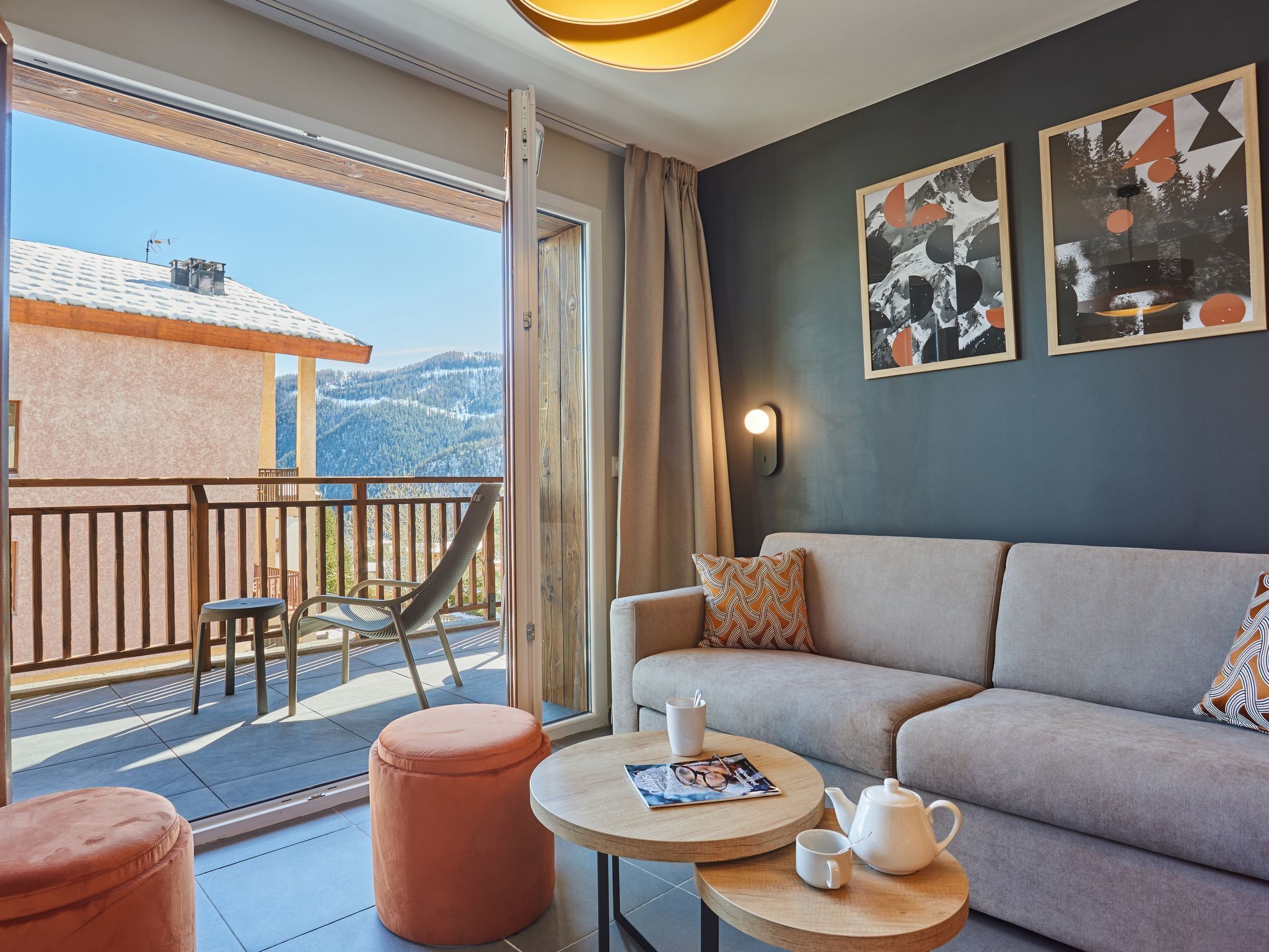 A modern living room with a sofa, ottoman, and round table with a teapot and magazine. A balcony with a view of mountains and a building is visible.