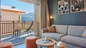 Un salon moderne avec un canapé, un pouf et une table ronde sur laquelle se trouvent une théière et un magazine. On aperçoit un balcon d’où l’on a une vue sur les montagnes et un immeuble.