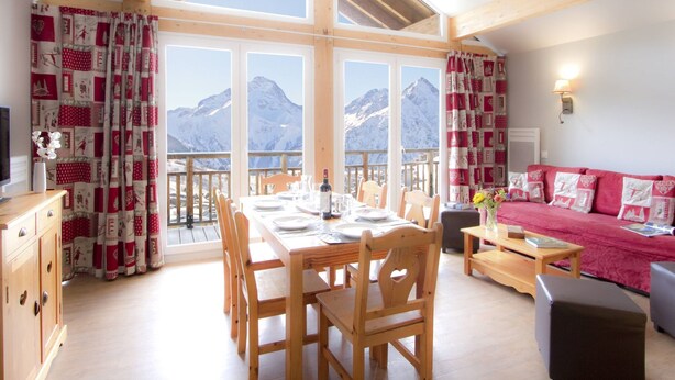 A cozy room with a wooden dining table, chairs, a red sofa, and a balcony with mountain views. - Odalys Référence L'Ours Blanc by Interhome (Les Deux Alpes)