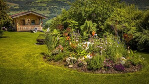 Une maison rustique en bois, avec un jardin devant, entourée d’une végétation luxuriante et avec des montagnes en arrière-plan.