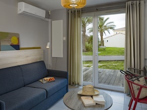 Ein modernes Zimmer mit einer Couch, einem runden Tisch mit Büchern und einem Hut, und Blick auf eine Palme sowie ein Haus durch eine Schiebetür.