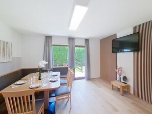 Uma área de refeições com uma mesa posta para quatro pessoas, uma televisão na parede e vista para um jardim pelas janelas.