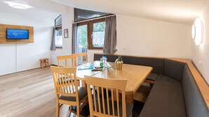 A dining area with a wooden table, four chairs, a sofa, a television mounted on the wall, and a window with curtains. - Alpenchalets by Interhome (Zell am See)
