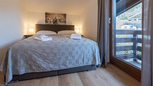 A hotel room with a bed, two bedside lamps, a painting on the wall, and a view of a balcony with buildings outside. - Alpenchalets by Interhome (Zell am See)