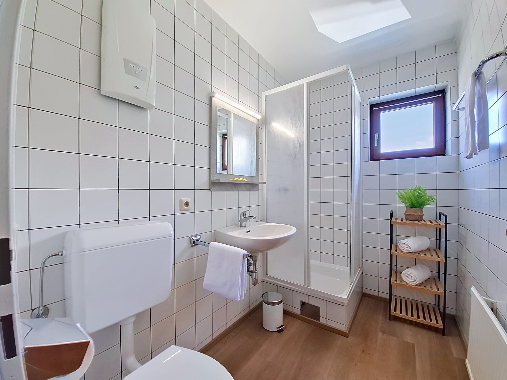 Une salle de bain avec des carreaux blancs, des toilettes, un lavabo, une douche, une fenêtre et une étagère avec des serviettes.