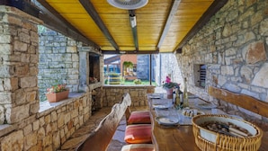 A rustic stone-walled room with a wooden bench, a table with plates and a bottle, and a ceiling fan. - Vacation home Bozac by Interhome (Pula/Vodnjan)