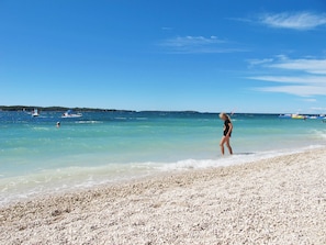 A beach with clear turquoise water, pebbly shore, and boats in the distance.