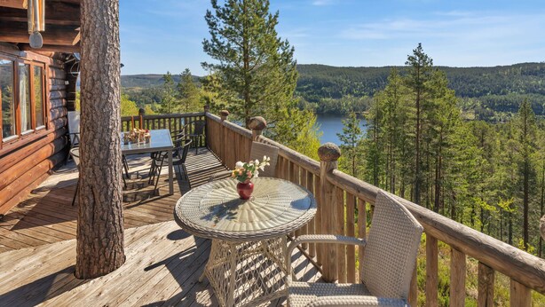 Une terrasse en bois avec une table et des chaises, offrant une vue sur une forêt et un lac.