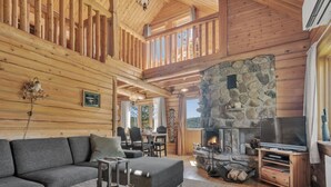 L’intérieur chaleureux d’un chalet avec une cheminée en pierre, un téléviseur posé sur un meuble en bois, un canapé et un coin repas avec des chaises.