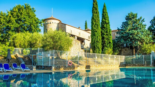 Una piscina con sillas de descanso, un edificio de piedra con una torre, y altos árboles.
