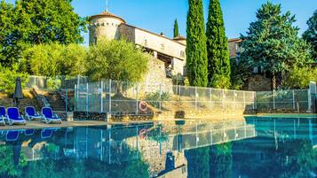 Une piscine avec des chaises longues, un bâtiment en pierre doté d’une tour et de grands arbres.