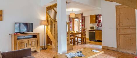 A living room with a sofa, a wooden coffee table, a TV on a wooden cabinet, a staircase, a dining area with a table and chairs, and a kitchen with wooden cabinets and a microwave.