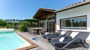 Un espace aménagé au bord de la piscine, avec une terrasse en bois, des chaises longues, une table avec des chaises et un grand parasol.