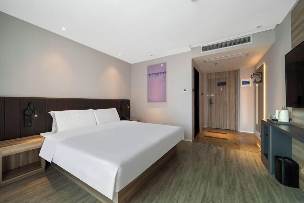 A modern hotel room with a large bed, a wooden headboard, a bedside table, a wall-mounted TV, and a bathroom with a wooden door.