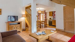 A living room with a sofa, a wooden coffee table, a TV on a wooden cabinet, a staircase, a dining area with a table and chairs, and a kitchen with wooden cabinets and a microwave. - La Cascade by Interhome (Vaujany)