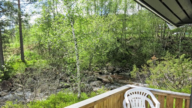 A wooden deck with a white plastic chair and a table, overlooking a rocky stream and dense greenery. - Thorabu by Interhome (Eikerapen)