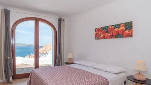 A bedroom with a bed, a nightstand, a lamp, a painting, and a view of the sea. - Hibiscus by Interhome (Palau)