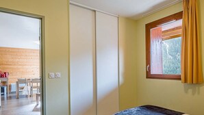 A bedroom with a bed, a window with curtains, and a view of a wooden wall and a dining area.