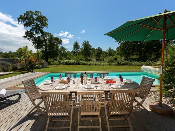 Uma área de refeições à beira da piscina, com um deck de madeira, uma mesa redonda para quatro pessoas e um grande guarda-sol.