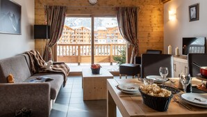 A cozy room with a wooden ceiling, a dining table set with plates, glasses, and a basket of snacks. A clock is visible on the wall, and there are curtains on the windows. - Le Lodge des Neiges 5* by Interhome (Tignes)