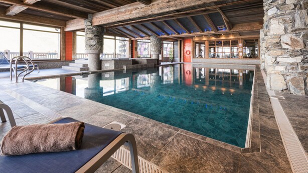 Una piscina interior con paredes de piedra y vigas de madera, una zona de estar con una toalla y una silla, y vistas al exterior.