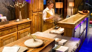 Una zona de bar con barra de madera, una mujer trabajando en la barra, un jarrón con flores y un menú sobre la mesa.