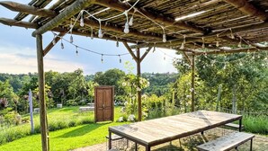 Een houten pergola buiten, met lichtjes, een stenen terras en uitzicht op een weelderige tuin en heuvels in de verte.