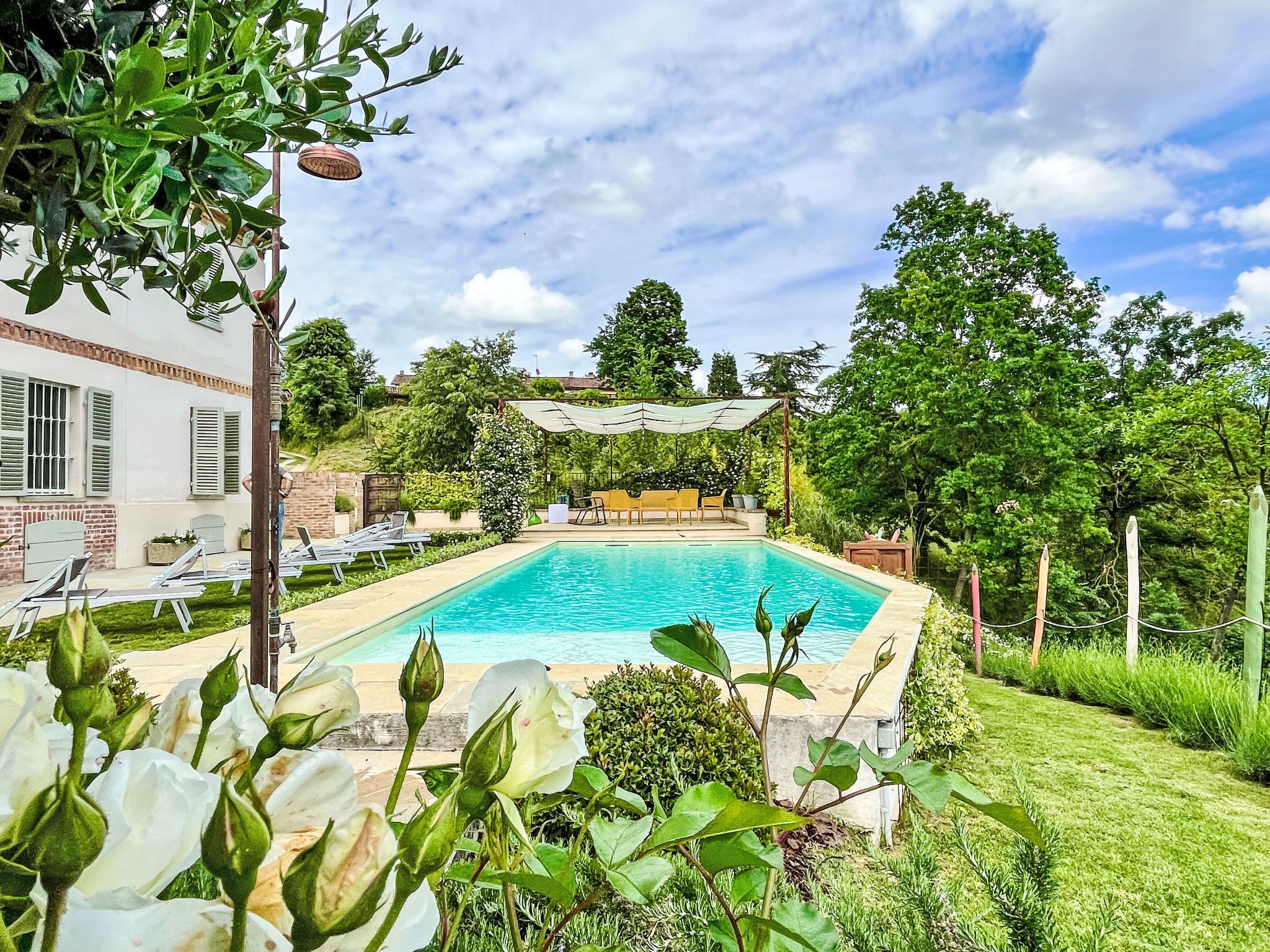 A swimming pool surrounded by greenery, with a building in the background and outdoor seating.