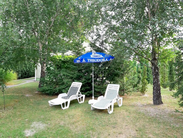 Two white lounge chairs under a blue umbrella with "THEODORA" written on it, surrounded by trees.