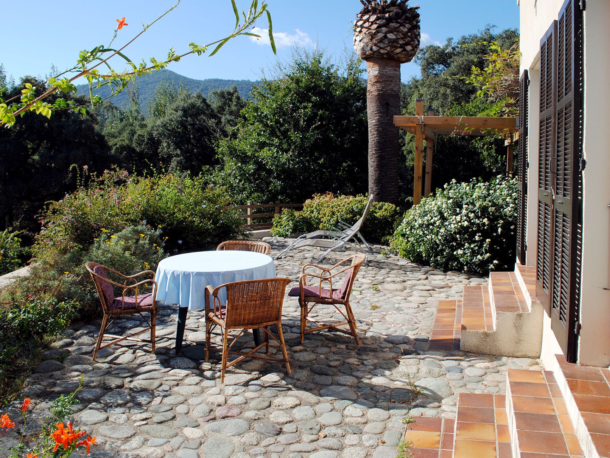 Eine Terrasse mit einem runden Tisch und Korbstühlen, umgeben von einem gepflasterten Weg und üppiger Bepflanzung.