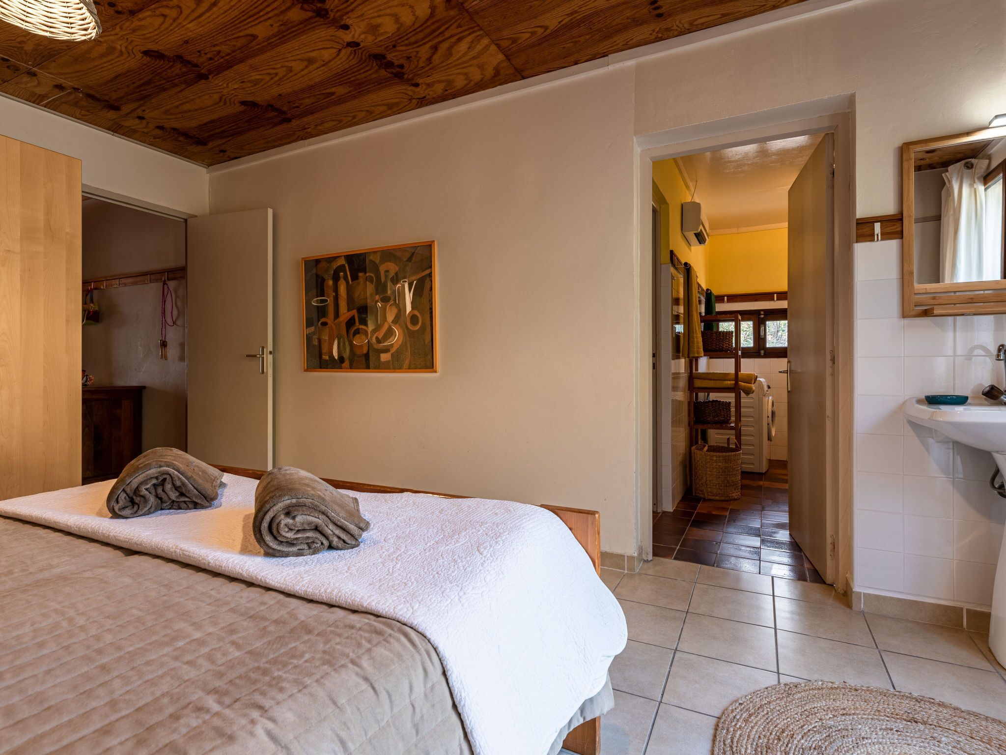 A bedroom with a bed, a painting, and a bathroom visible through an open door.