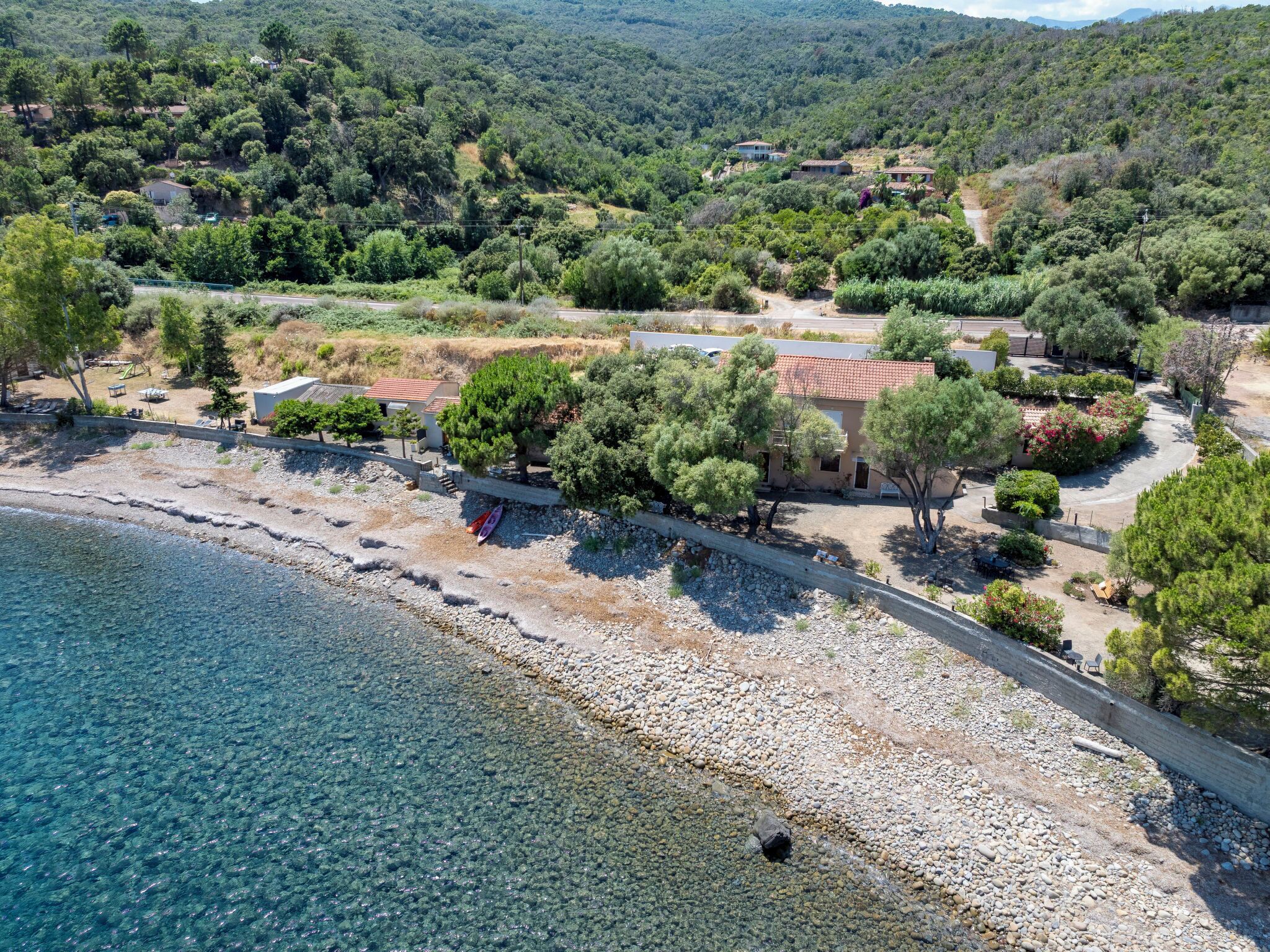 Une propriété située sur la côte, avec une plage, une eau cristalline et un chemin menant à une maison entourée d’arbres et de végétation.