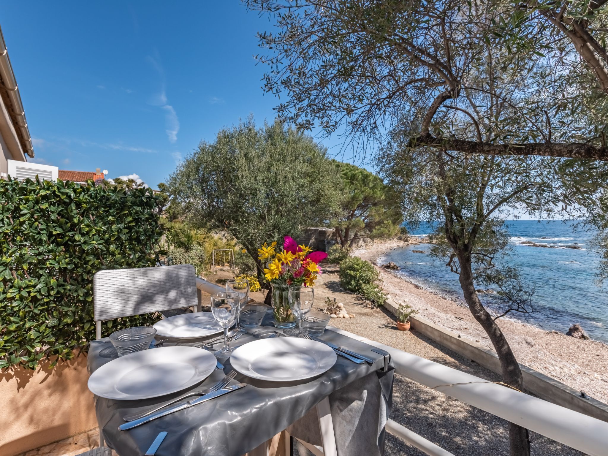 An outdoor dining table set with plates, cutlery, and a vase of flowers, overlooking a coastal area with trees and a view of the sea.