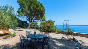 A round metal table with chairs, a bench, and a small statue in a garden with a view of the sea. - Vacation home Polpu by Interhome (Solenzara)