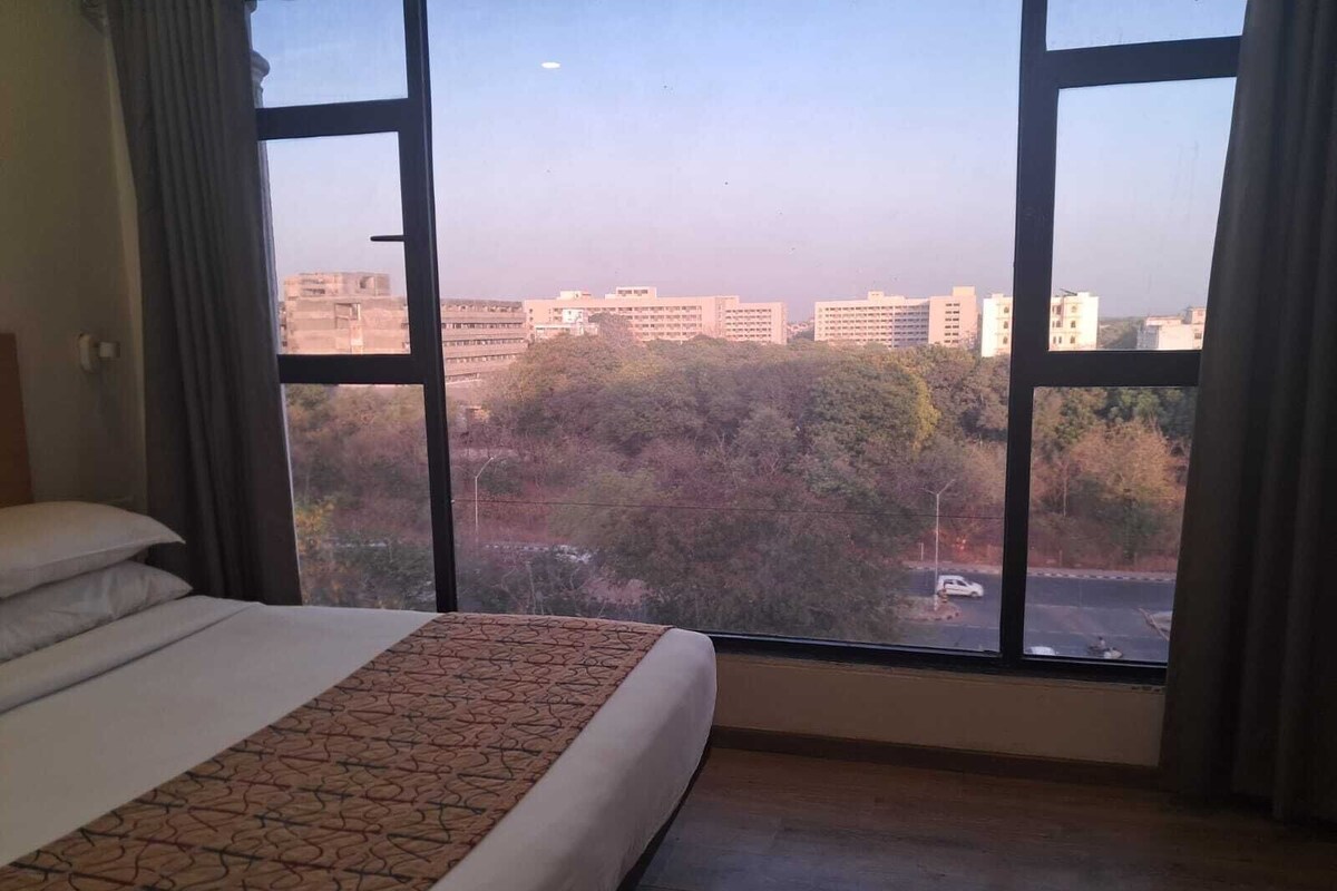 A hotel room with a large bed, a window with a view of buildings and trees, and a curtain.