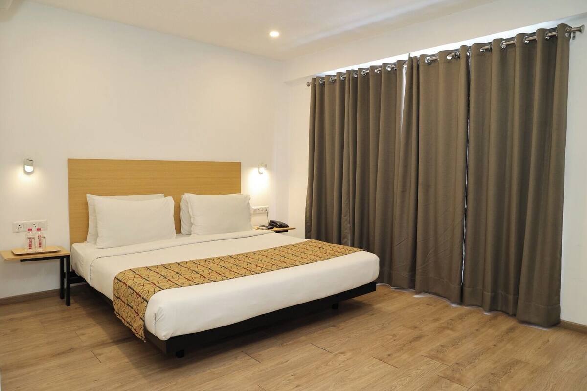 A hotel room with a wooden headboard bed, white bedding, a patterned throw, a bedside table with a phone, and a curtain.