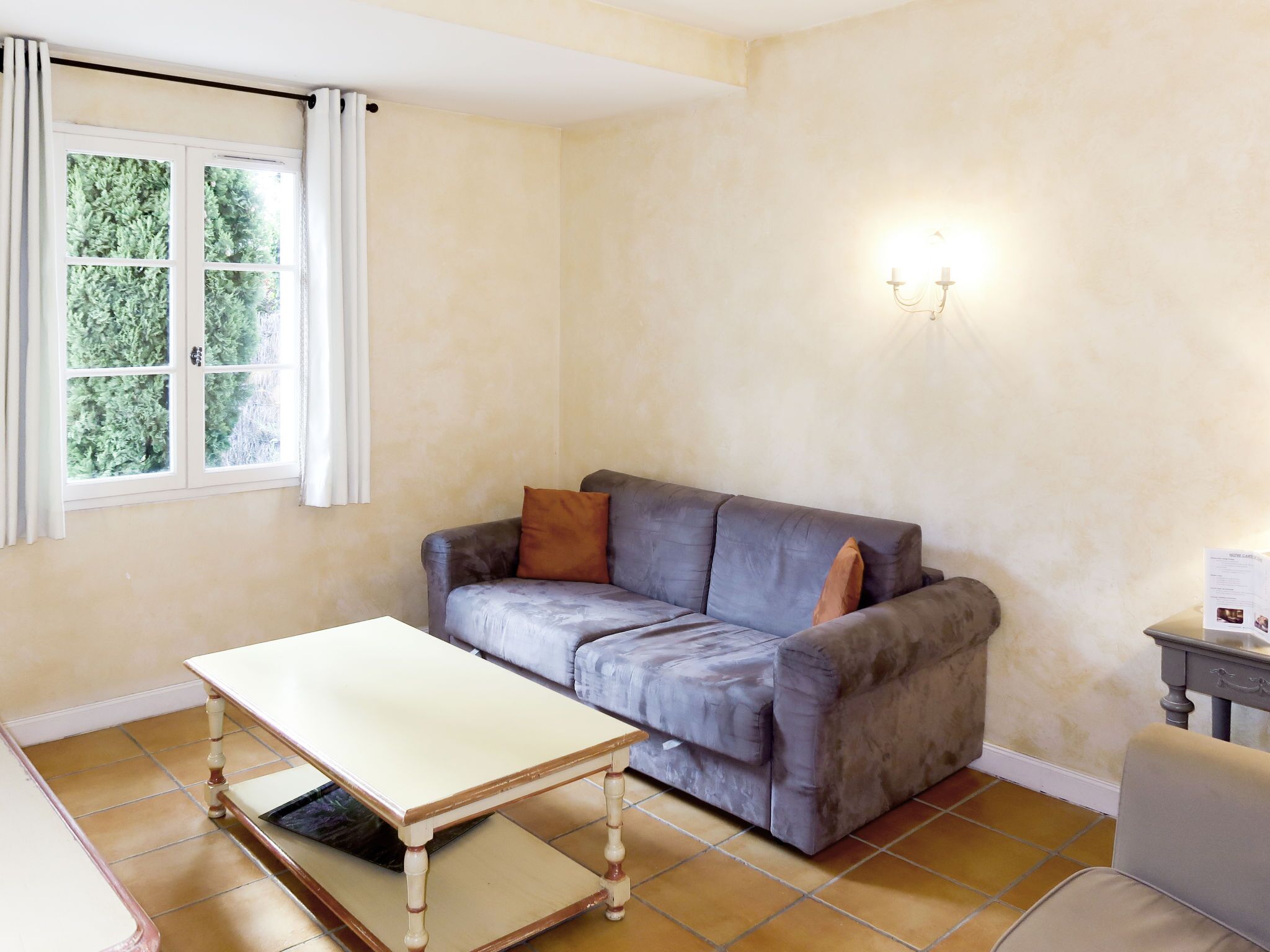 A room with a sofa, a table, a window with curtains, and a wall-mounted light fixture.