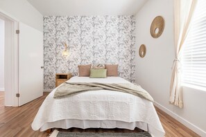 Een slaapkamer met bloemenbehang, een bed met een witte sprei, een bedlamp en een raam met blinds.