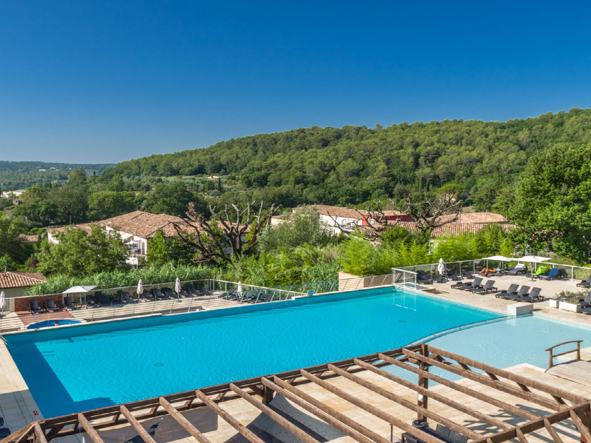 A swimming pool with sun loungers and a wooden deck, surrounded by lush greenery and houses with red-tiled roofs.