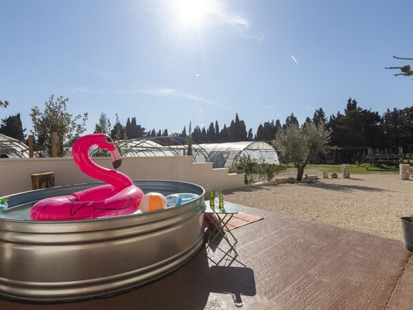 A hot tub with a pink flamingo float, surrounded by a garden with potted plants and a greenhouse in the background.
