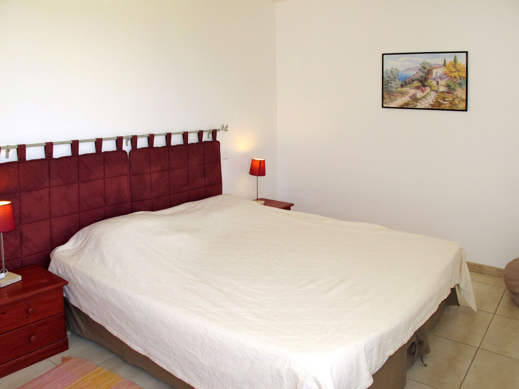 A neatly made bed with a red headboard, a bedside table with a lamp, and a framed picture on the wall.